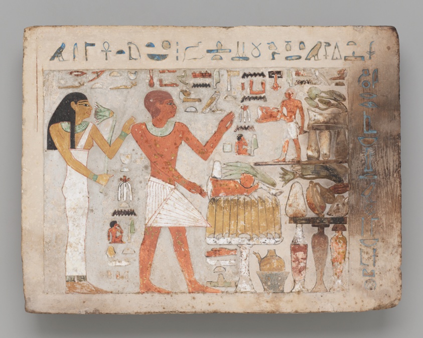 A rectangular stone slab is painted with two large figures with medium-dark skin in profile, a man and a woman. Surrounding them are various food items, as well as a smaller male figure holding a large piece of meat. Across the top are hieroglyphics.
