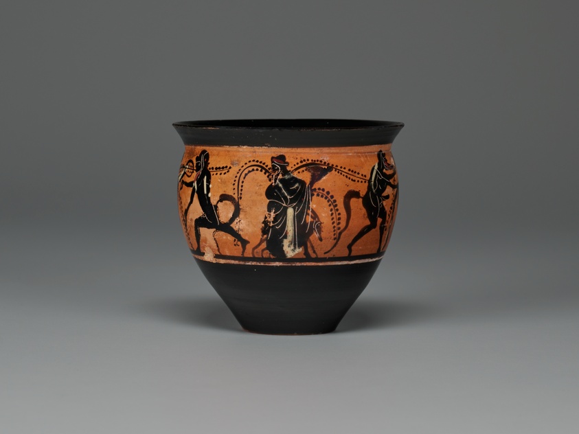 Black terracotta vessel, narrow at the bottom and wider at top, with a short lip, featuring a wide red band in the middle. This band depicts in black a procession of figures surrounded by vines, including a person on a donkey.