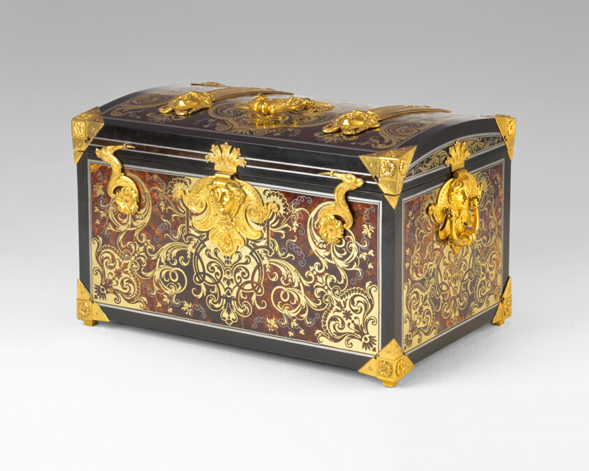 Ornate wooden box with golden metallic corners and embellishments, painted golden flourishes throughout. Its gold metallic clasp is embossed with a human face.