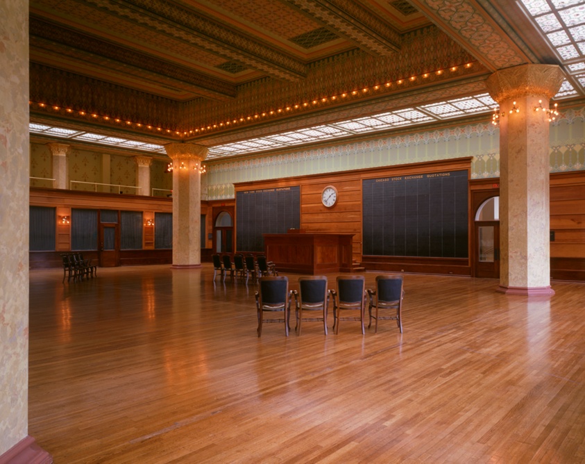 A grand, double-height room with wood paneling, stenciled wall patterns, four hexagonal columns, and an interior balcony with guardrail at left.