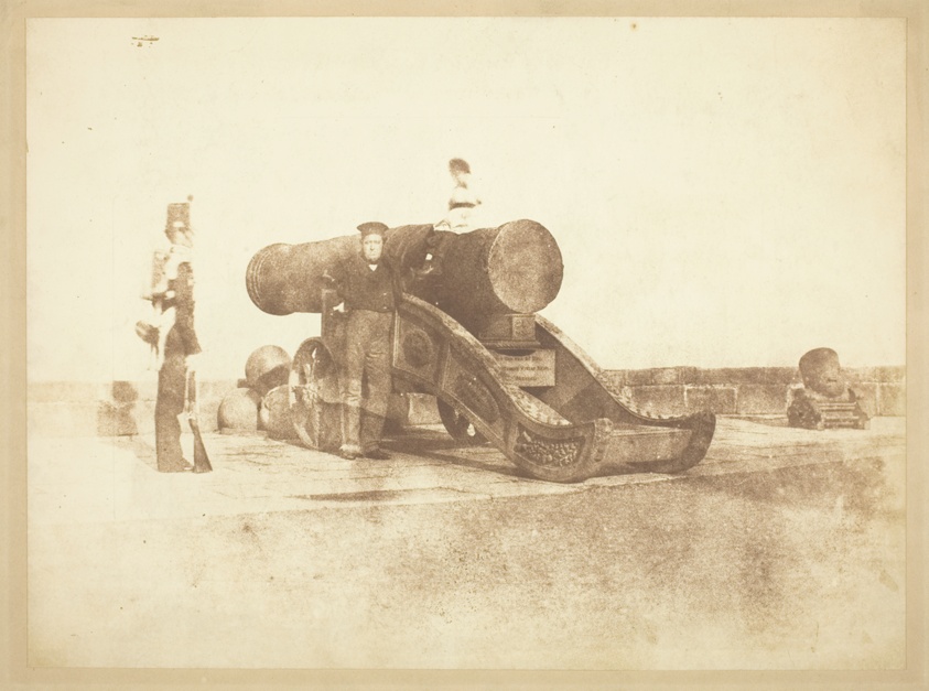 Sepia-colored, blurry photo of a grim-faced man in a flat-topped hat posing next to a cannon. A small, blurred figure sits on top of the cannon while another figure, in uniform with a rifle at his side, stands at left.
