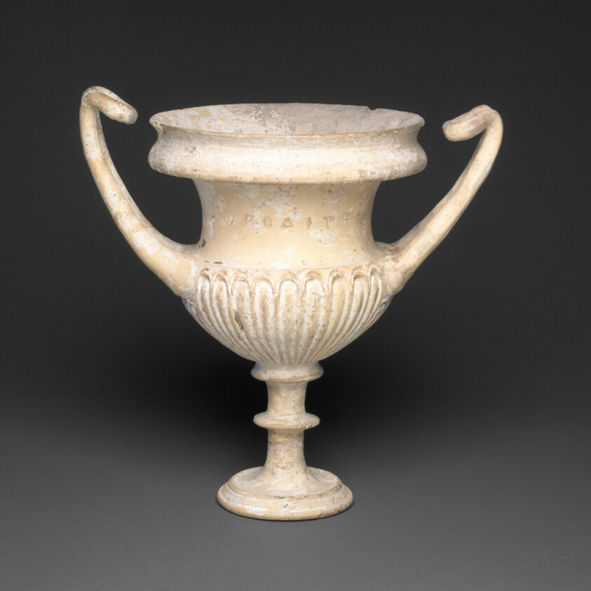 Cream-colored cup with a small round foot and a thin stem. Two thin arms flare up and out from mid-cup in a "V" shape and bend inward without reconnecting to the cup. The cup's opening tapers in before flaring out again at the lip. Greek letters are inscribed on the cup between the arms and lip.
