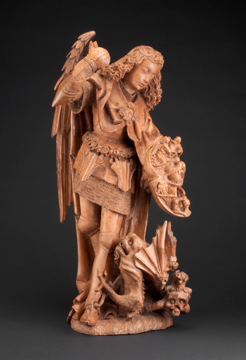 Light wood sculpture of angel with shield looking towards devil at feet.