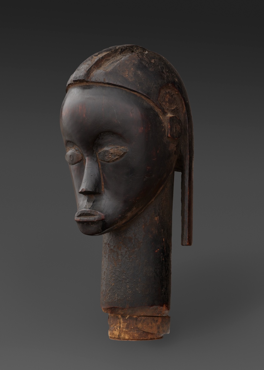 A dark-brown painted sculpture with hints of orange showing through of a slim face with a long neck. The lips are drawn in a pout, the cheeks sunken, the hair pulled back in a swirl pattern and falling down in a slim column.