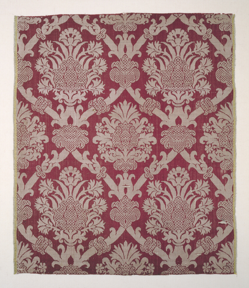 Textile with a cream-colored floral motif on red fabric, vines arranged in repeating diamond shapes, a large flourish or blossom within each one.