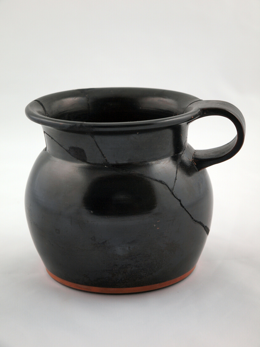 Black glazed terracotta vessel with a short, curved handle on one side. Visible cracks indicate fragmentary repair.