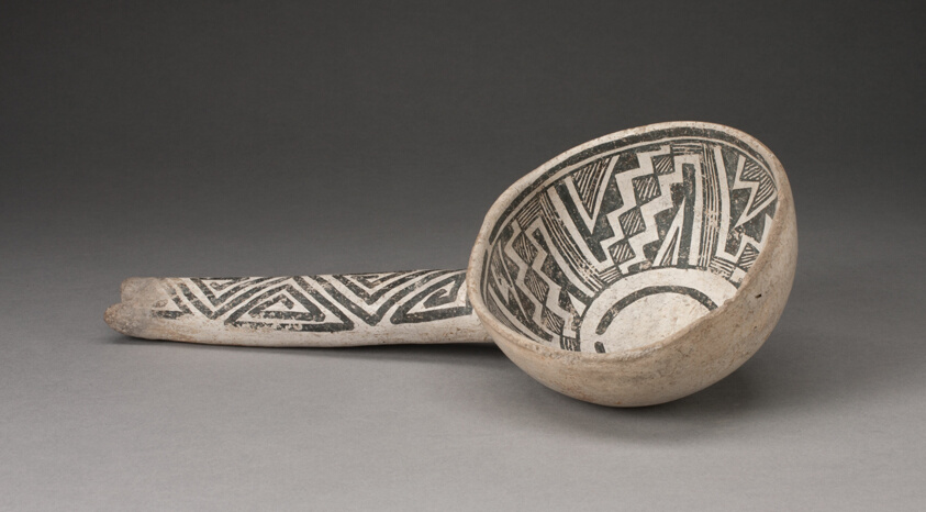 A ceramic vessel with a long handle, its bone-white surface decorated with black geometric designs.