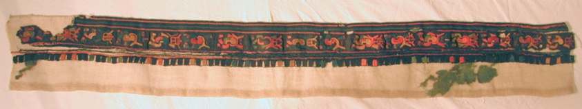 Textile border fragment in blue, green, and coral, with stripes at top, figures in the middle, and square fringe at bottom.