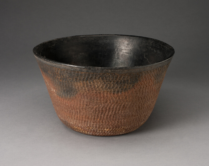 Conical-shaped bowl with sides angled outward, a smooth black interior, and a bumpy exterior that is black along the rim and graduates to a reddish-brown color.