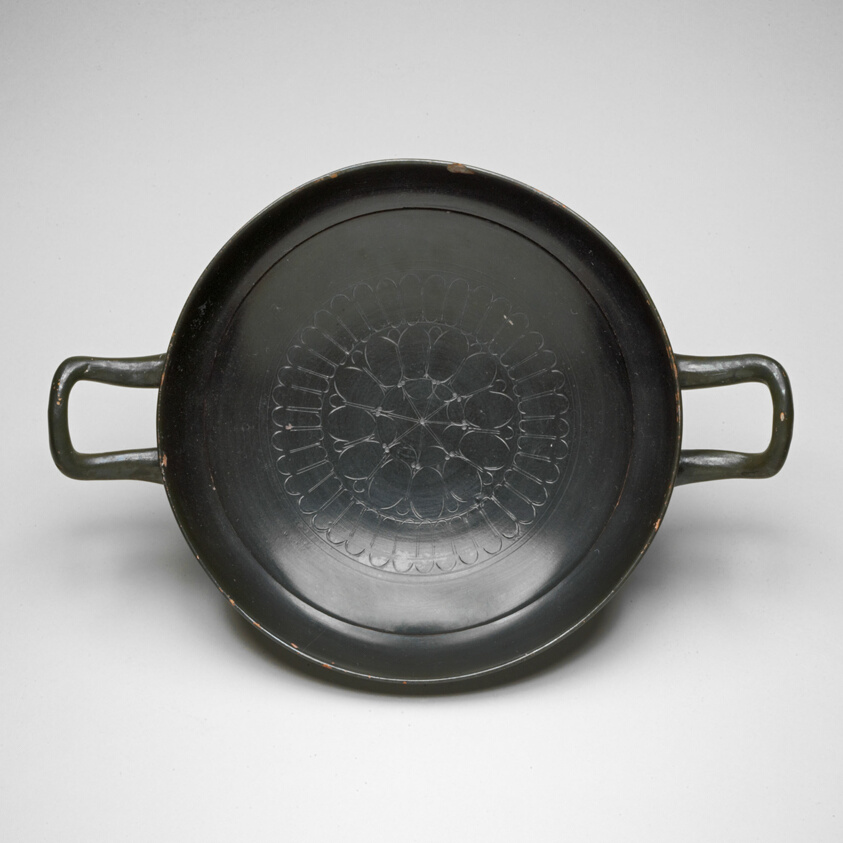 Shallow terracotta pan-like cup with black glaze, incised floral decoration at bottom, and handles on either side.