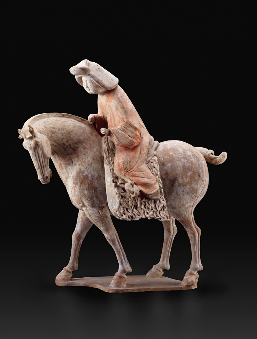 A female figure with a round face in a full orange robe is bent almost wearily as she rides a horse. White pigment has worn away on the horse, and orange has worn from her robe, revealing a terracotta tone.