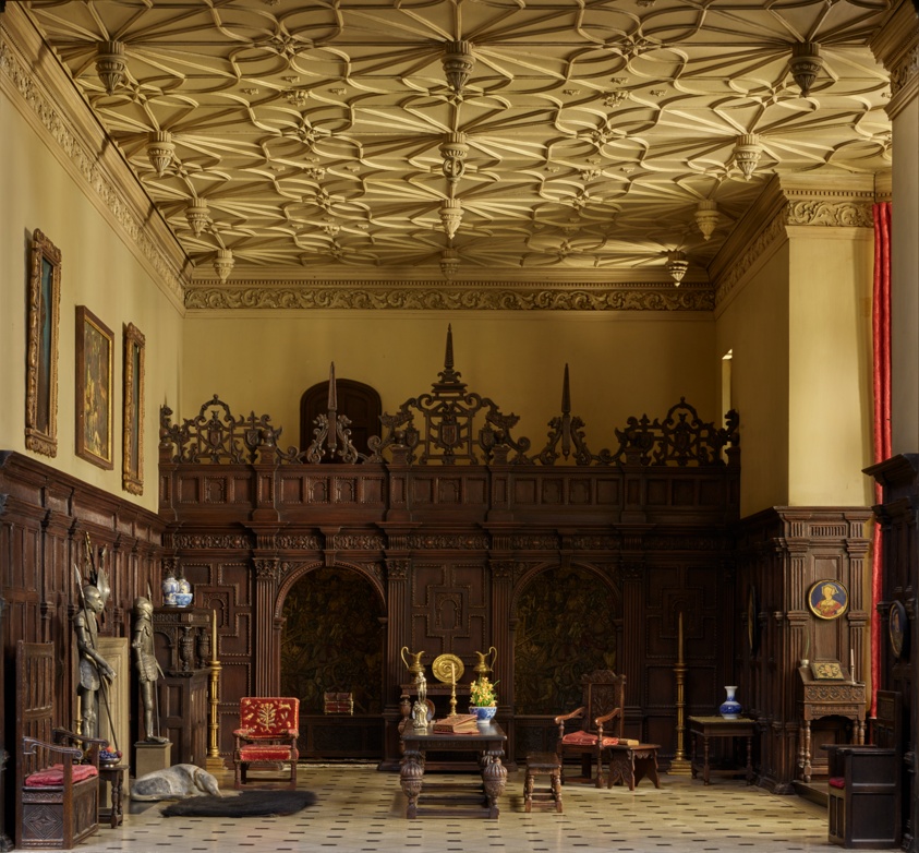 To-scale great room with a double-height ceiling. The bottom half of each wall is clad in elaborately carved wooden panels. The right wall features a tall bay window, the left a fireplace flanked by suits of armor. A long, rustic table with bench is centered in the room.