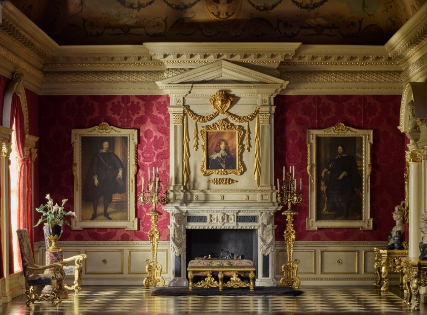 Miniature of an ornate room with red walls and white and gold trim, a large fireplace at center, and black and white checkered flooring. Two armchairs flank a window on the left side. Three large painted portraits in golden frames hang on the back wall. There is a small table directly in front of the fireplace.