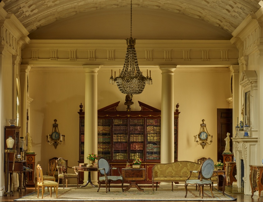 Miniature room with cream-colored walls with inset columns on the side walls and two columns at the back framing a dark wooden bookshelf. A large crystal chandelier hangs from a curved ceiling patterned with geometric designs in relief. A beige floral rug fills the floor, upon which a yellow couch and other upholstered wood furniture are unevenly spaced. On the right wall is an ornate fireplace with a mantle framing a painting of a landscape.
