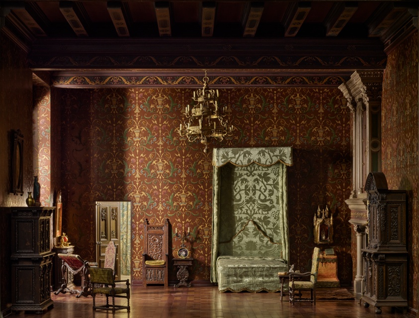 Miniature of a very dark, ornate bedroom with a partially canopied green bed at center, brown parquet flooring, and deep brown and golden wallpaper. A gold chandelier hangs in the center, and two dark-brown cupboards sit on either side of the room. On the right is also a white and brown fireplace.
