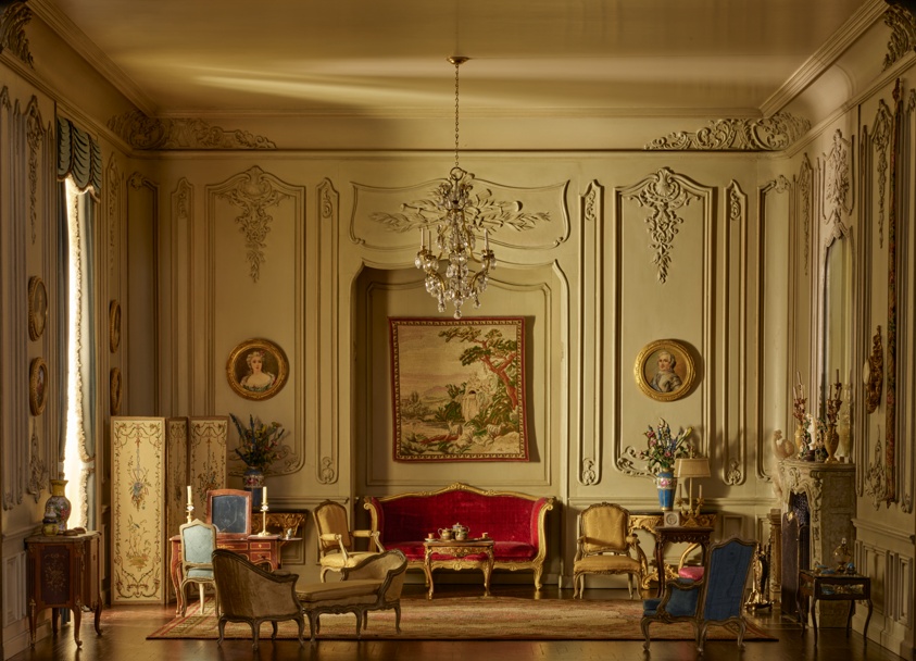 Ornate interior with cream walls and dark wood floors. At left is a large window with blue curtains; at right is a fireplace with tall mirror. Gilded upholstered furniture and decorative elements, including numerous framed portraits, fill the room. A crystal chandelier hangs from the ceiling.