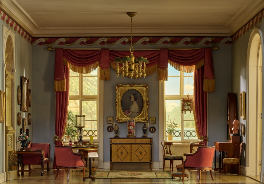 Replica of a sitting room with pale-blue walls and a wave-patterned wallpaper border. Heavy red velvet curtains on the center wall frame two tall windows, between which hangs a painting of a woman in a gold frame. A beige stone fireplace is at left, an open arched doorway at right.