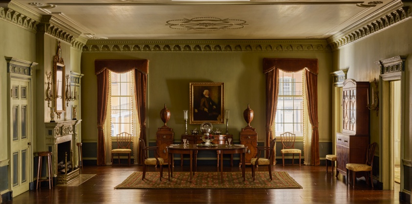 Miniature dining room with mint-green walls and two large windows on the back wall, between which hangs a portrait of a Revolutionary-era man flanked by two vases. A formal wooden table with two chairs sits in the room's center, a fireplace with a mirror on the mantle and two tall stools at left.