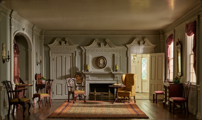 Miniature scale model of a large, furnished white-walled room with ornamental moulding, wooden flooring, upholstered wooden chairs, and a large decorative carpet in the floor's center.