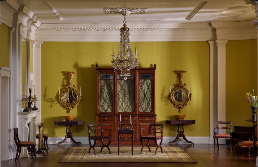 A miniature room with yellow walls, inset pillars, and an ornate cabinet centered on the far wall. At room's center, a large chandelier hangs over a small table with two upholstered chairs.