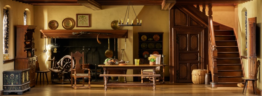 Scale miniature of a rustic but refined kitchen with a varnished wooden table at center and one chair. A dark wooden chest sits beneath the window at left, a spinning wheel in the left back corner. In the right corner is an ornately carved door adjacent to a staircase. Metal plates line a wide hearth.