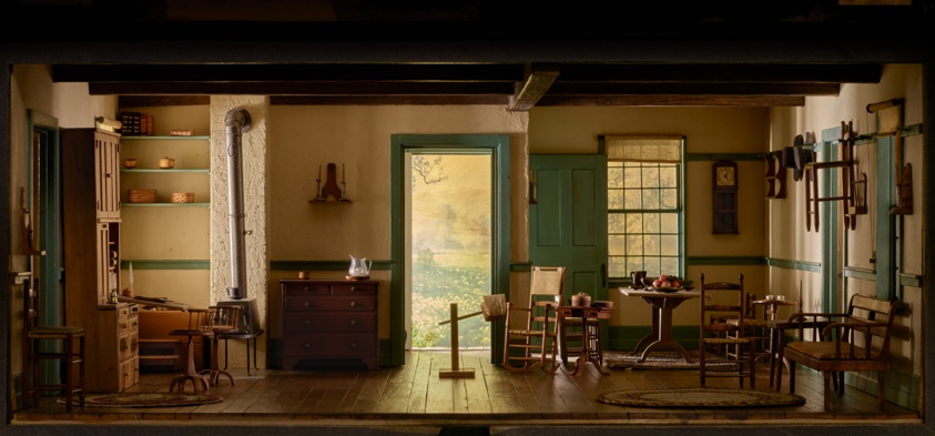 Miniature room featuring wide-plank hardwood floors, round hooked rugs, simple wooden furnishings, and a wood-burning stove. The window frames and door frames are painted green, while the walls are a light butter yellow.