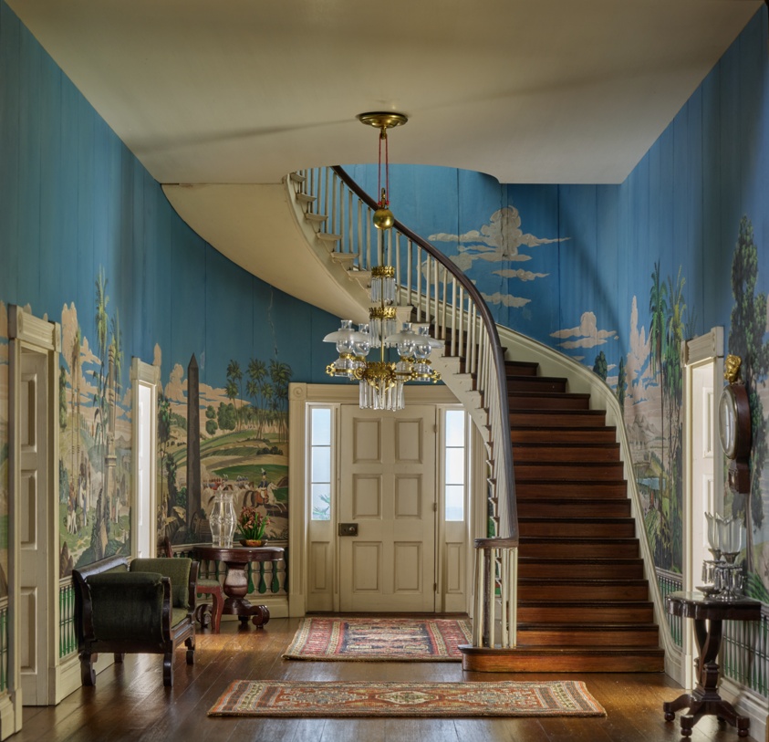 Miniature room featuring walls decorated with a tropical landscape, a spiral staircase, a crystal chandelier, and two rugs.