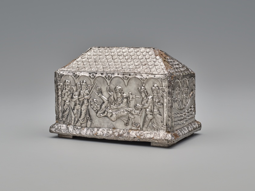 Silver-toned reliquary in the shape of a long rectangle with an upper resembling a pitched roof. In relief on its side, armed figures surround a falling figure in distress as a single figure attacks him.
