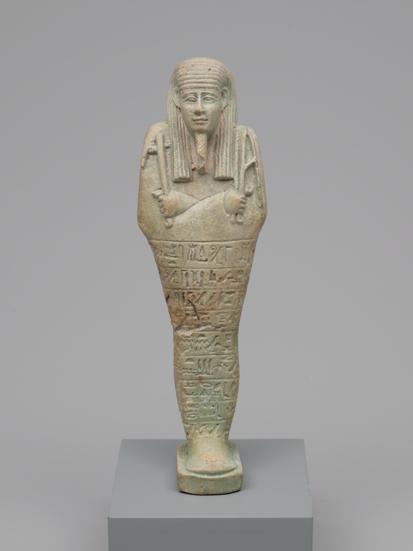 A full-body figure of a human carved in what looks to be light-colored stone. The figure wears an ancient Egyptian style headdress, its arms are crossed holding symbolic instruments, and the body is covered with carved symbols.