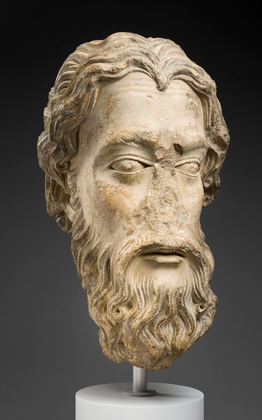 Sculpture in light stone of the head of a bearded man with wavy hair and wide eyes, missing a nose.