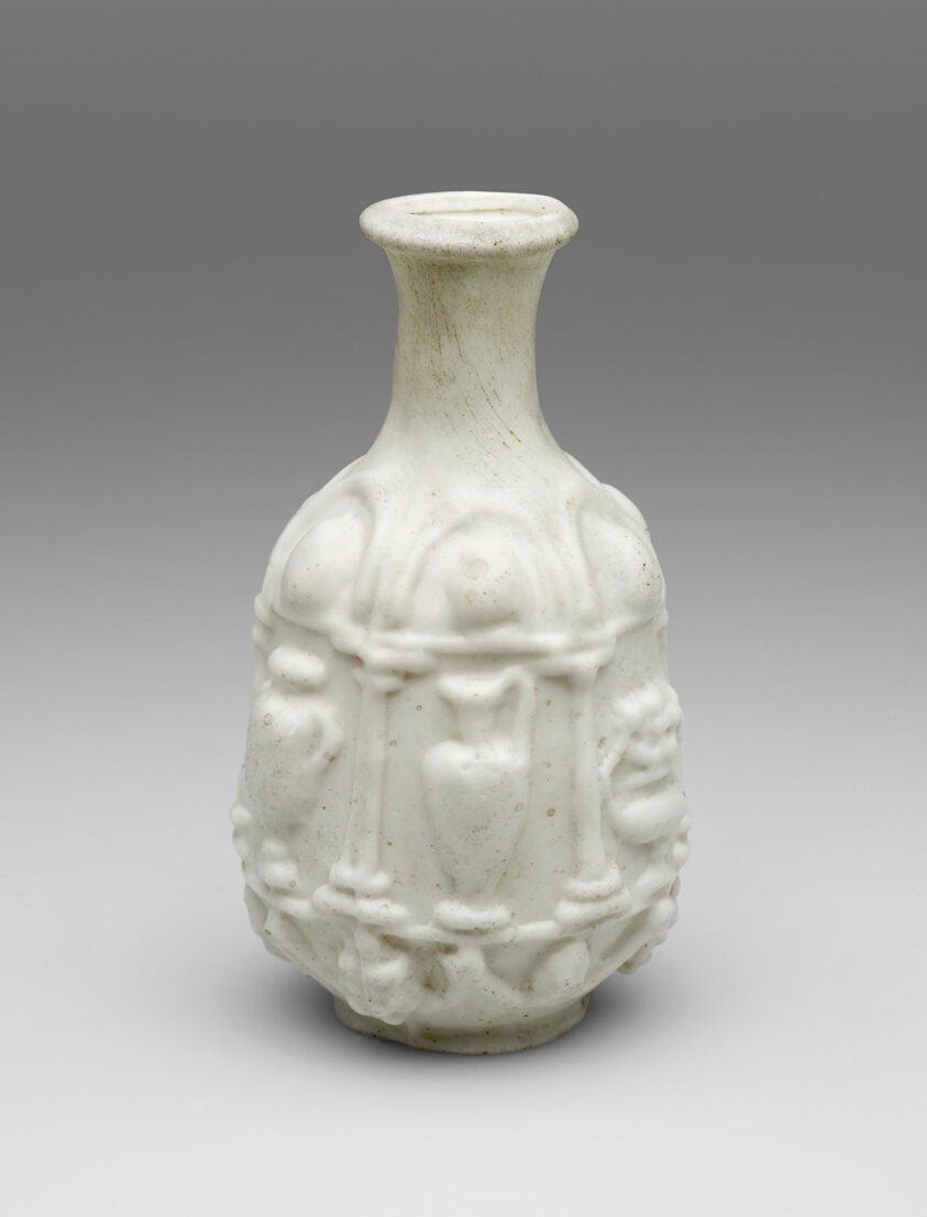 A white glass bottle with a narrow neck decorated with reliefs of columns and vases or pitchers.