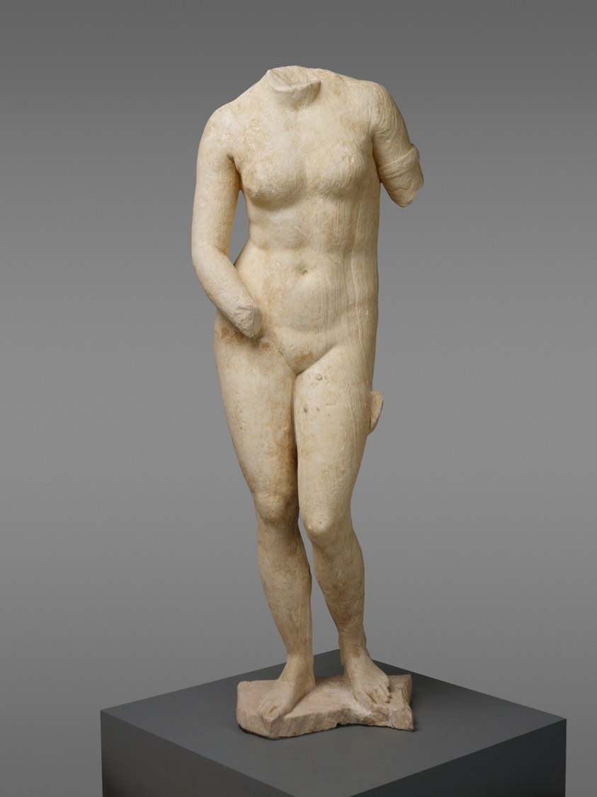 White and beige stone statue of a female nude standing in contrapposto position, with weight on the right leg. The head of the statue is missing. The left arm has been broken off just past the shoulder, and the left hand is also absent.