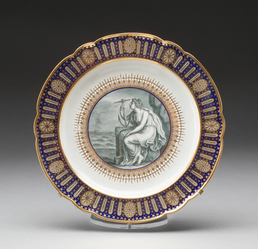 Porcelain plate with scalloped rim decorated with a border of royal blue and gold. The center is a black and white enamel depicting a female figure holding an anchor and seated on a rocky shore.