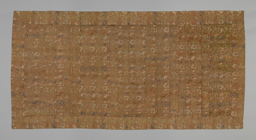 Rectangular, mostly brown textile made up of many small fabric squares of the same pattern, featuring flora and fauna in green, blue, and cream.