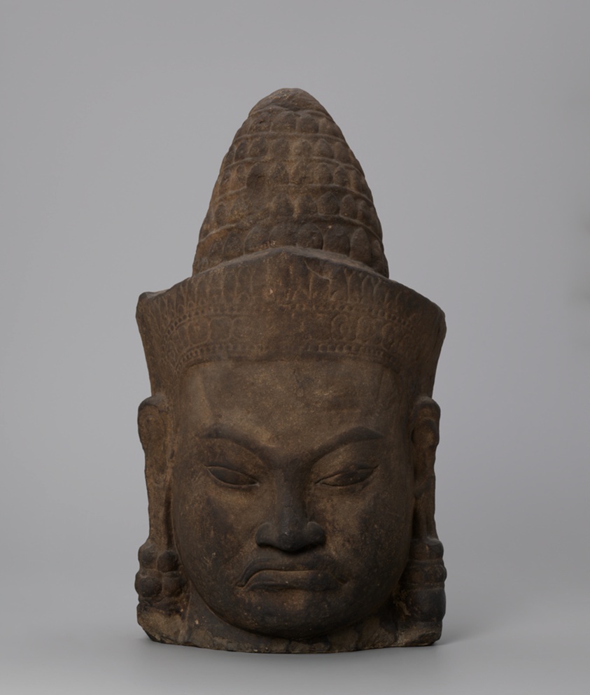 Tan stone carving of a head, features weathered darker, headwear with geometric pattern.