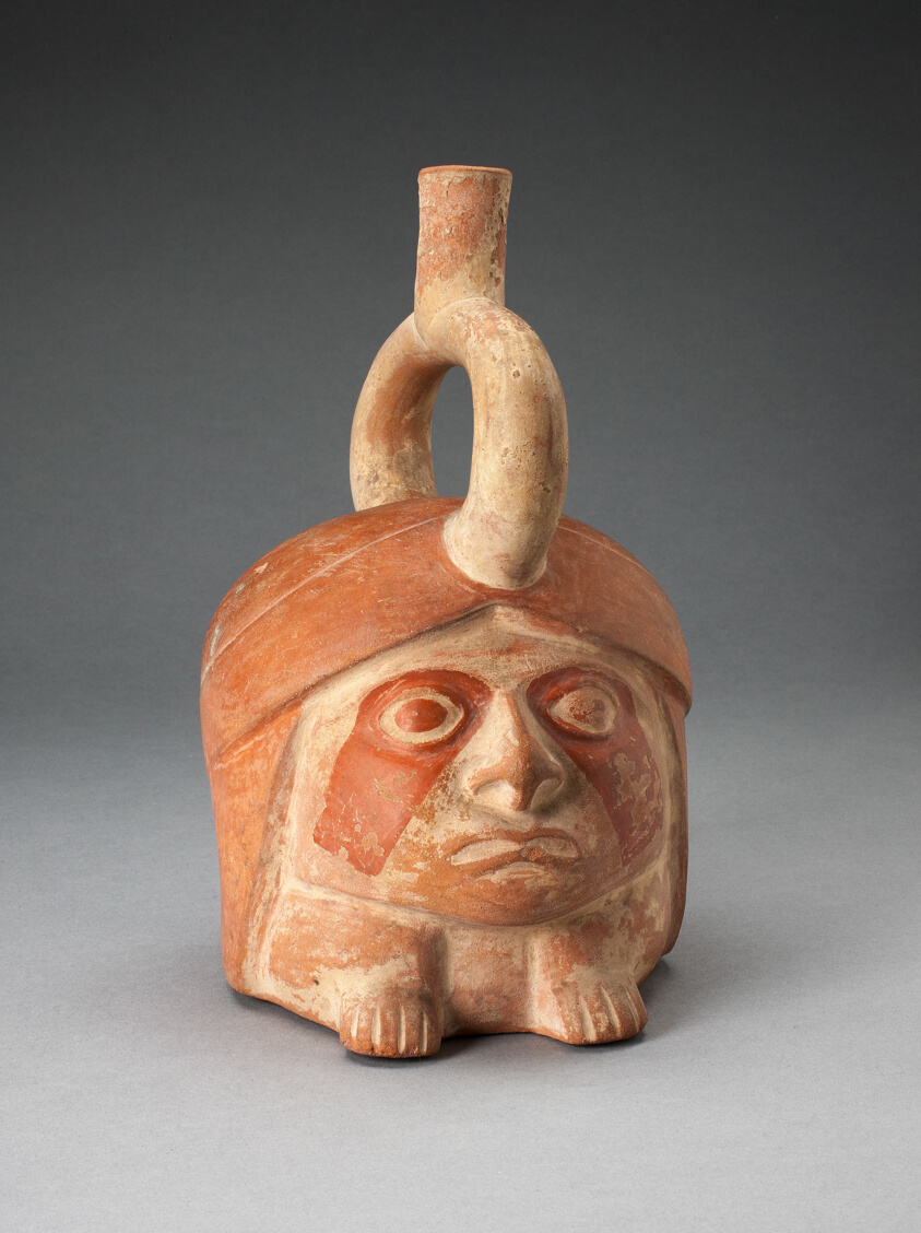 A head-shaped tan and red-orange ceramic vessel with a round, expressionless face atop two squat feet, darker markings around the eyes and down the cheeks.