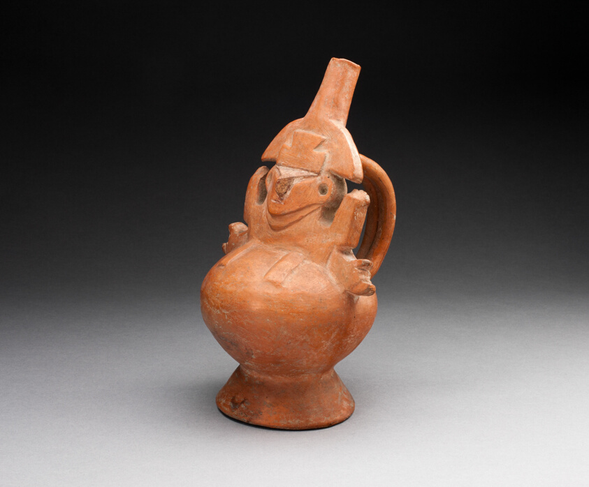 An orange-red ceramic vessel in the shape of an abstract figure with wide-set eyes and short arms, a handle at back.